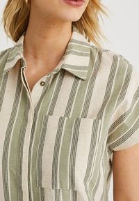 Chemisier à manches courtes avec des rayures verticales vertes et crème, doté d'une poche poitrine et d'un col. Tissu texturé.