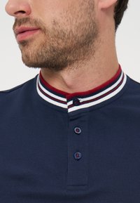 Άνδρας με γενειάδα που φοράει μπλε πουκάμισο με γιακά με κόκκινες, λευκές και μπλε ρίγες και τρία σκουρομπλε κουμπιά στην πατιλέτα.