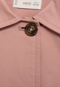 Vaaleanpunainen kauluspaita, jossa on ruskea nappi ja kontrastivärinen tikkaus. Liukas kangas, jossa on siisti viimeistely ja näkyvä napinreikä.