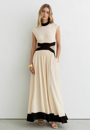 Kobieta ubrana w beżową, bezrękawnikową suknię maxi z czarnymi wycięciami w talii, czarnym obszyciem na dole, czarnym kołnierzem, złotymi bransoletkami i beżowymi sandałami.