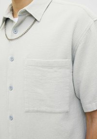 Camisa de botones de mangas cortas en un gris claro, hecha de una tela texturizada, con un bolsillo en el pecho y botones azul claro. Se aprecia un detalle de cadena plateada.