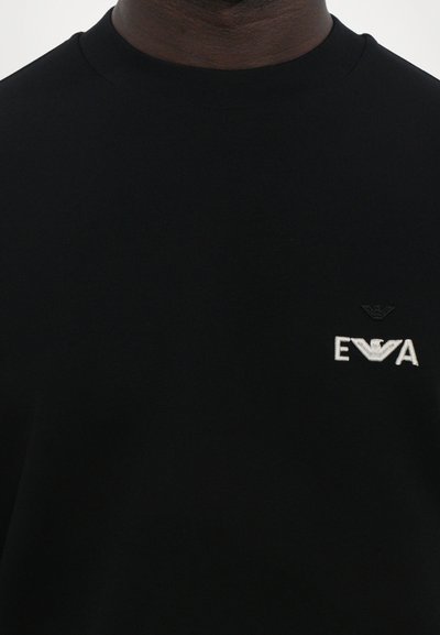 T-shirt en coton noir à col rond avec un petit logo brodé blanc sur la poitrine gauche, offrant une texture lisse et une coupe classique.