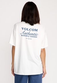Biała bawełniana koszulka z niebieskim nadrukiem na plecach, który brzmi „VOLCOM Authentic Durable Goods”, o luźnym kroju i krótkich rękawach.
