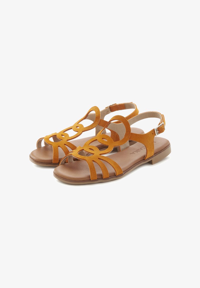Tan suede sandals featuring a strappy design with circular accents and an open toe. Flat sole and ankle strap with a buckle closure.