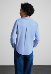 Camicia azzurra a maniche lunghe con yoke posteriore e texture morbida, abbinata a pantaloni in denim blu scuro. La vista posteriore mostra un orlo stondato.