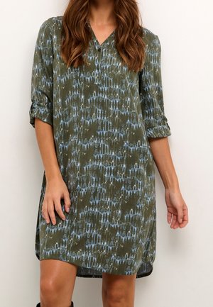Femme portant une robe mi-longue vert olive avec un motif abstrait bleu et des manches trois-quarts retroussées, debout devant un fond blanc.
