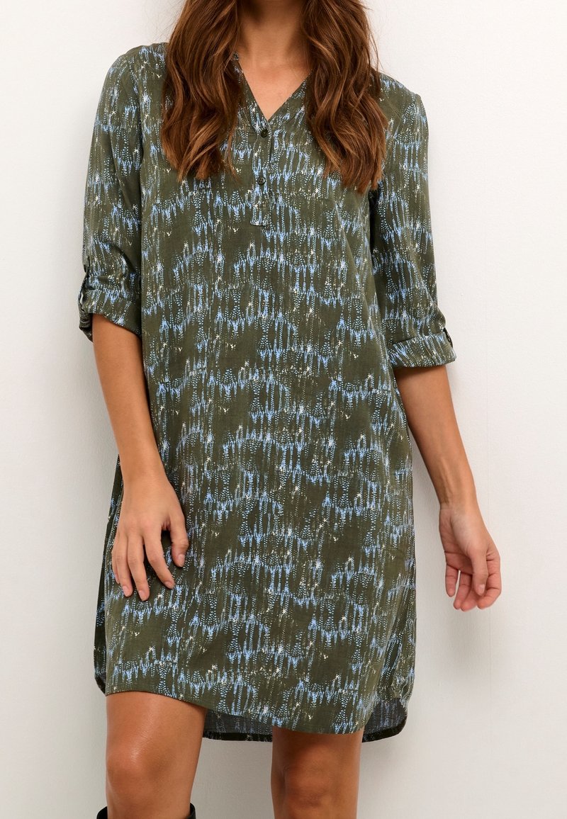 Femme portant une robe mi-longue vert olive avec un motif abstrait bleu et des manches trois-quarts retroussées, debout devant un fond blanc.