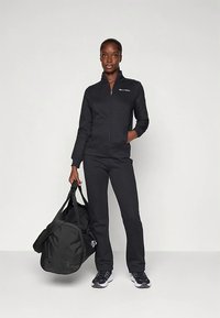 Kvinne i svart jakke med glidelås og bukser som holder en stor svart treningsbag, stående mot en ensfarget hvit bakgrunn.