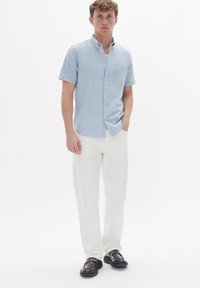 Camicia azzurro chiaro a maniche corte con bottoni e taschino sul petto, abbinata a pantaloni bianchi e scarpe marrone scuro. Tessuto morbido, vestibilità casual.