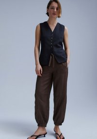 Woman stands wearing sleeveless black buttoned vest, loose brown drawstring pants with elastic cuffs, and black open-toe heeled sandals on gray background.