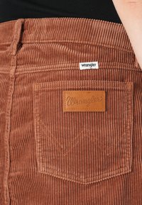 Brown corduroy pants featuring a back pocket with a leather patch and orange stitching, branded with "Wrangler" on a tag.