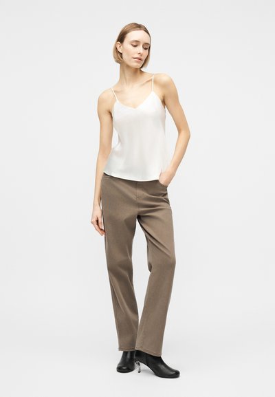 Femme debout, une main dans la poche, portant un haut caraco blanc, un pantalon marron et des bottines noires, sur un fond clair uni.