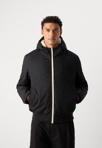 Giacca nera isolata con cappuccio, dotata di una cerniera crema, foderata in peluche e con polsini a costine. Design semplice con tasche laterali.