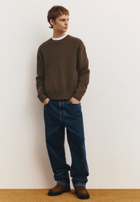 Barna bordázott pulóver kerek nyakkivágással, laza kék farmernadrággal és barna csizmával, egyszínű háttér előtt.