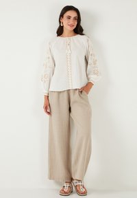 Blusa bianca con ricami floreali beige e scollatura con coulisse, abbinata a pantaloni larghi beige chiaro e sandali decorativi.
