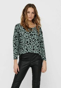 Pull en imprimé léopard vert et noir avec un large col et des manches longues, porté avec un pantalon en simili cuir noir. Texture tricot douce.