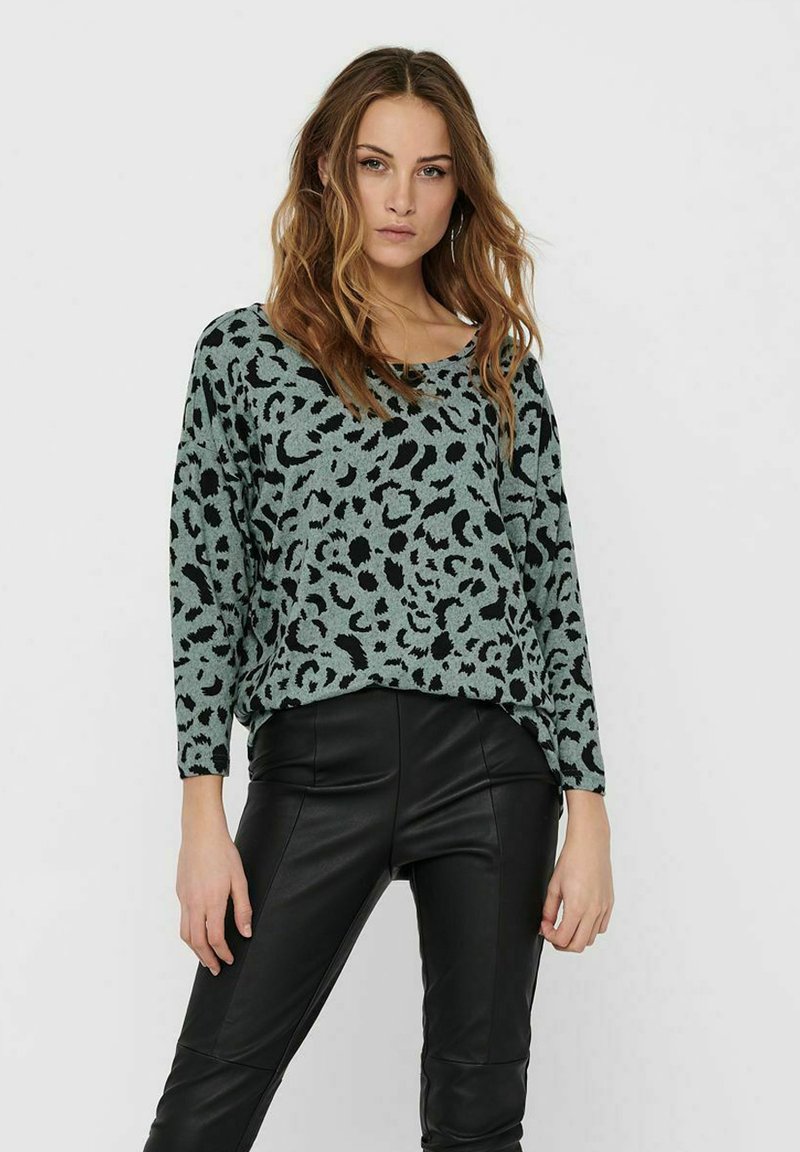 Pull en imprimé léopard vert et noir avec un large col et des manches longues, porté avec un pantalon en simili cuir noir. Texture tricot douce.