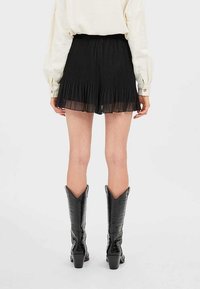 Pantalones cortos negros con pliegues y un dobladillo superpuesto translúcido, combinados con botas de vaquero negras hasta la rodilla. El material texturizado añade profundidad al diseño.