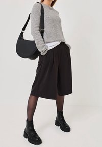 Personne portant un pull gris, un sous-vêtement blanc, un pantalon culotte noir, des collants noirs transparents, des bottines noires à lacets et un sac à épaule noir.