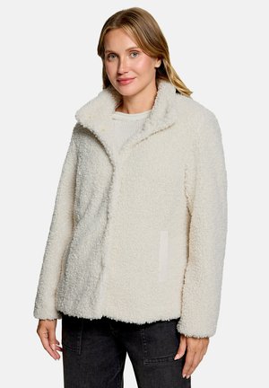 Kvinde med blondt hår bærer en hvid, fluffy fleecejakke over en let sweater og sorte bukser, stående mod en ensfarvet baggrund.