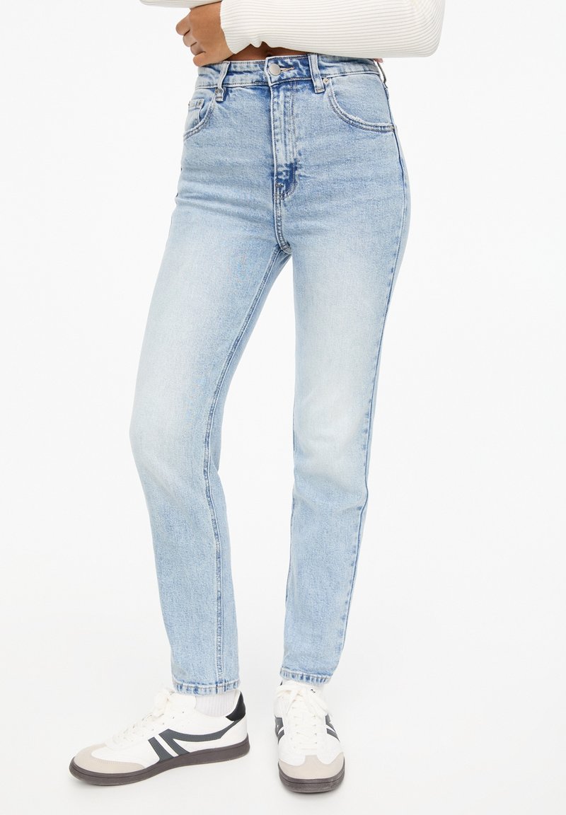 Femme portant un jean taille haute bleu clair et des baskets blanches avec des bandes noires, debout, les bras croisés sur un haut blanc.