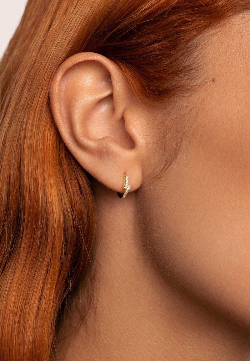 Orecchino a cerchio in oro con design curvato, decorato con piccoli gemmi scintillanti, esposto su uno sfondo di pelle liscia e chiara.