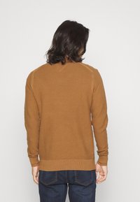 Maglione a coste marrone con maniche lunghe, scollo a giro e orlo aderente, abbinato a jeans blu scuro. La trama è prominente ed uniforme.
