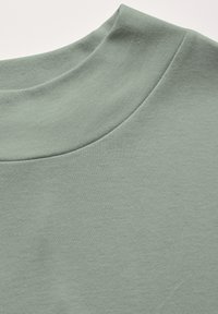 Světle zelená, strukturovaná látka s vysokým, zaobleným výstřihem. Hladký, měkký materiál s minimálním prošíváním.