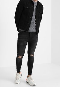 Svart denimjacka över en grå stickad tröja, ihop med svarta slitna skinny jeans och vita sneakers med blå och röda detaljer.