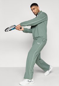 Πράσινη φόρμα από μαλακό ύφασμα, με λευκές λεπτομέρειες. Ένα άτομο κρατάει ρακέτα τένις, φοράει λευκά αθλητικά παπούτσια.