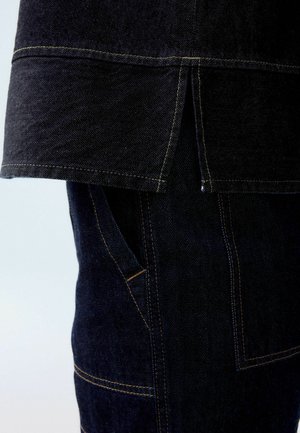 Denim-vaate tummansinisellä värillä. Ominaisuuksiin kuuluu kontrastin ompelulla viimeistelty helma ja sivutaskut, jotka esittelevät tekstuuriompeleista kangasta ja räätälöityä muotoilua.