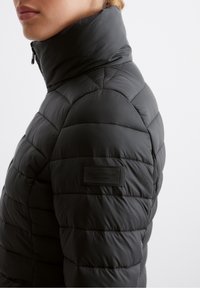 Sort dunjakke med høj krave, prydet med quiltet tekstur og et mærke på ærmet. Glat, letvægtsmateriale.