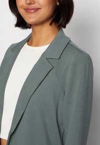 Blazer costine turchese con rever a lancia e design strutturato, abbinato a una camicia chiara e aderente. Focus su texture e contrasto di colore.