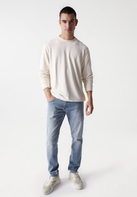 Camicia beige a maniche lunghe con collo a giro, abbinata a jeans azzurri chiari e sneakers color crema. Fit casual, design minimalista e texture morbida.