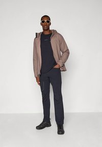 Svetlo rjava jopa s kapuco čez temno modro majico, kombinirana s temnimi hlačami in črnimi supergami. Modni dodatki vključujejo bele sončna očala.