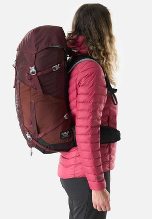 Personne portant une doudoune rouge et un pantalon noir, avec un grand sac à dos de randonnée bordeaux muni de plusieurs sangles et boucles.