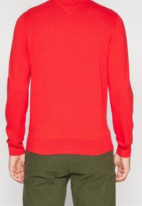Pull rouge à manches longues, avec poignets et ourlet côtelés. Col en V. Porté avec un pantalon vert olive, affichant un look simple et ajusté.