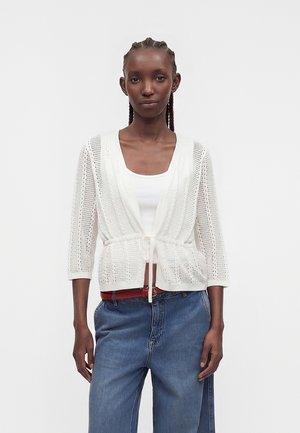 Femme avec des tresses portant un cardigan blanc en maille ouverte noué à la taille, un débardeur blanc, un jean bleu ample et une ceinture rouge.