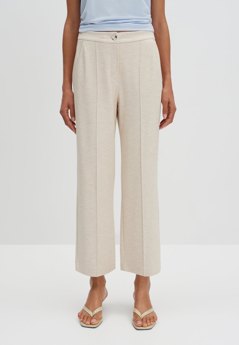 Beige wide-leg trousers with a textured finish, featuring pleats and a button closure at the waist. Worn with beige strappy sandals.