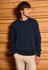 Marineblaue Pullover mit Rundhalsausschnitt aus einem glatten Strickstoff, mit langen Ärmeln und gerippten Bündchen. Kleines Logo-Detail auf der Brust.