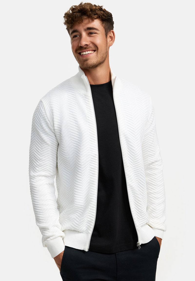 Weiße Zip-Jacke mit strukturiertem Fischgrätmuster, geripptem Kragen und Bündchen, kombiniert mit einem schwarzen Hemd. Verfügt über seitliche Taschen.