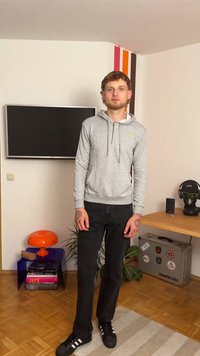 Sivkasta kapuco z zadrgo, s sprednjim žepom in kapuco, črne kavbojke ter črne športne čevlje z belimi črtami. Notranjost prostora vsebuje televizor in okrasne predmete.