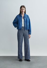 Femme debout, main dans la poche, portant une veste en jean bleue, un haut blanc, un pantalon large texturé bleu et des chaussures sombres sur un fond uni.