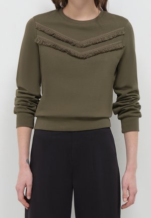 Femme portant un pull à manches longues vert olive avec deux bandes chevron texturées sur la poitrine et un pantalon noir, bras détendus sur les côtés.