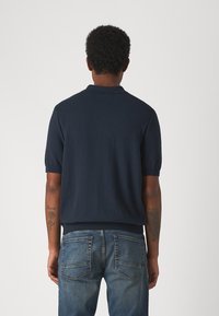 Polo en tricot marine à manches courtes, avec col et poignets côtelés, porté avec un jean en denim bleu. Tatouage visible sur l'avant-bras gauche.