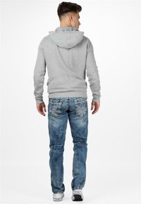 Felpa grigia con cappuccio e cordoncino, abbinata a jeans in denim blu con toppe sbiadite e design classico a cinque tasche. Sneakers bianche visibili.