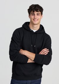 Felpa nera con cappuccio, tasca frontale, coulisse, polsini e orlo a costine. Indossata con jeans scuri su uno sfondo neutro.
