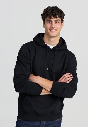 Felpa nera con cappuccio, tasca frontale, coulisse, polsini e orlo a costine. Indossata con jeans scuri su uno sfondo neutro.