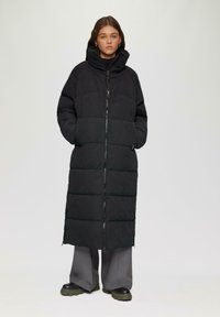 Lange zwarte pufferjas met hoge kraag, ritssluiting en twee zijzakken. Heeft een gewatteerd patroon en een licht oversized pasvorm.