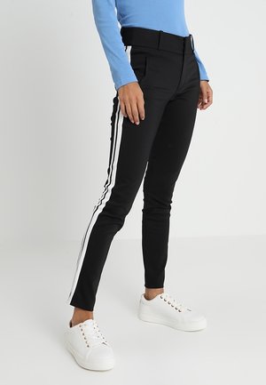 Person trägt schwarze schmale Hose mit weißen Seitenstreifen, blaues Langarmshirt und weiße Sneakers, steht auf weißem Boden.